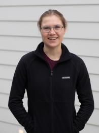 A white woman with glasses and pulled back hair is standing smiling in front of a wall. 